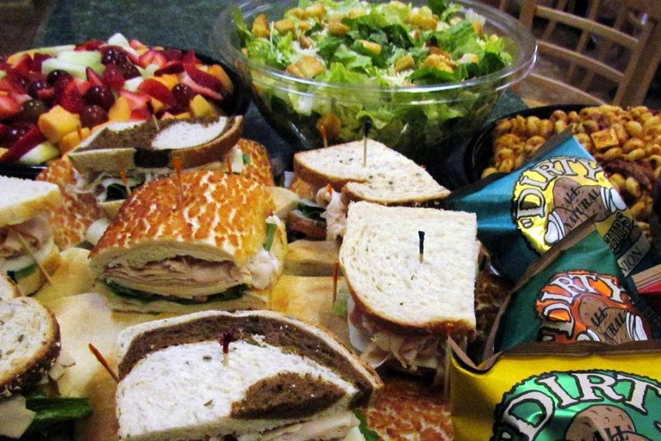 A catering spread: sandwiches, big salads, fresh fruit, and pasta